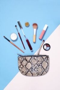 A flat lay of vibrant makeup items arranged around a stylish snakeskin cosmetic bag on a blue and beige background.