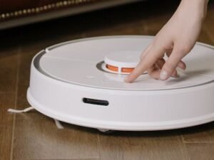 Close-up of a hand pressing a button on a robotic vacuum cleaner on a wooden floor.