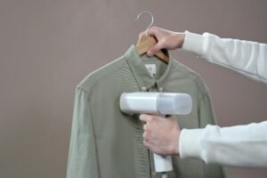 Hands steaming a green shirt with a portable steamer against a grey background.